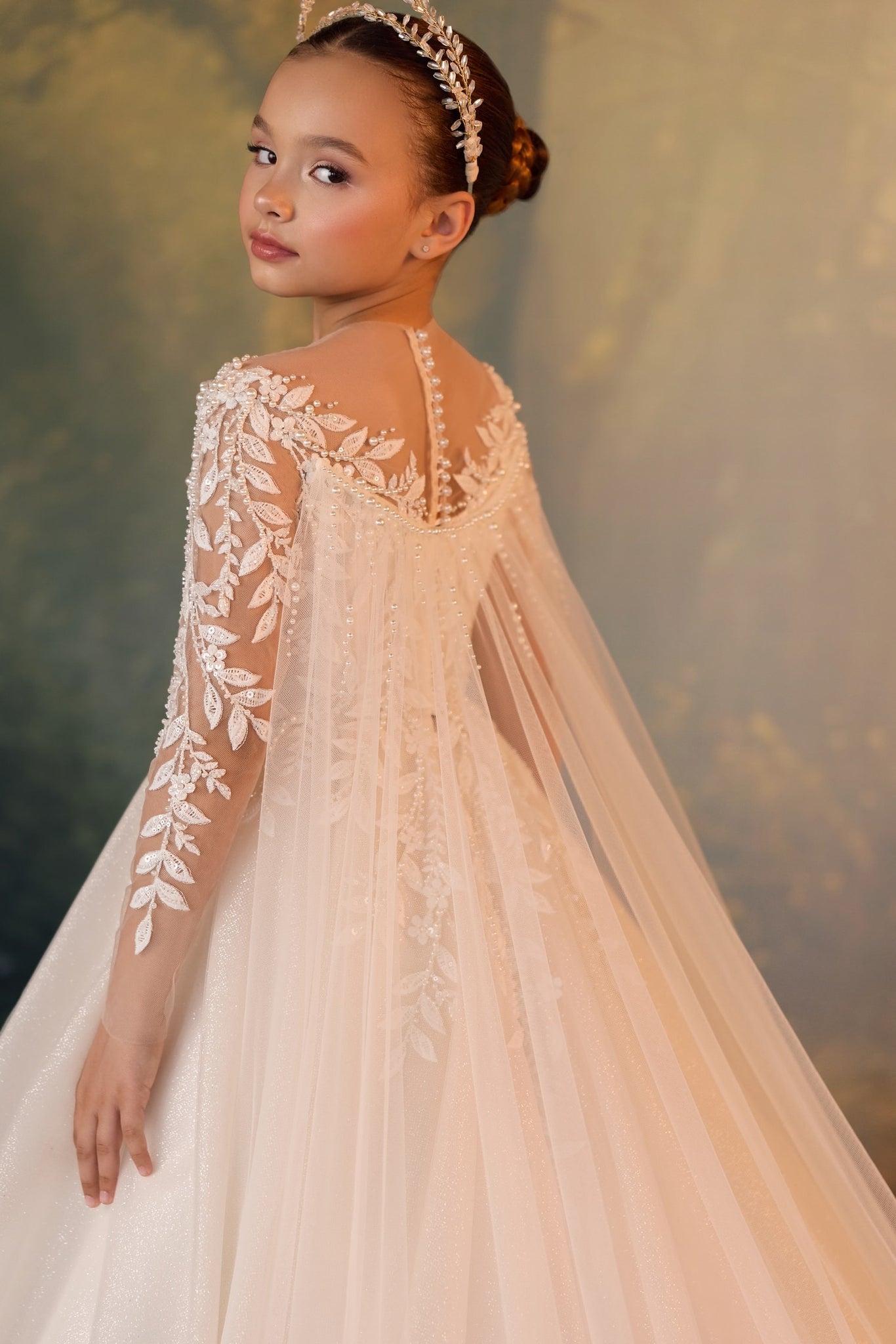 Girl wearing a white lace gown with long sheer sleeves and a capelet with pearl accents