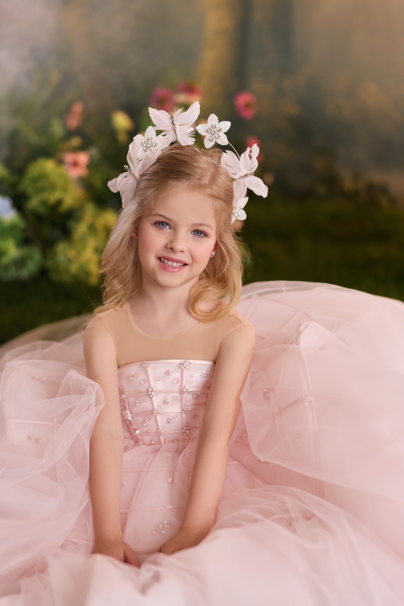A young girl with blonde hair wearing a white floral crown sits in a voluminous light pink ball gown with a strapless bodice. The background is a soft, blurred garden setting.