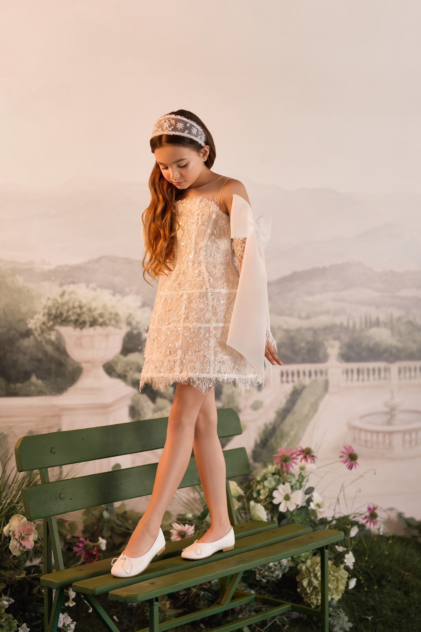 Girl wearing a white lace dress and headband standing on a green garden bench
