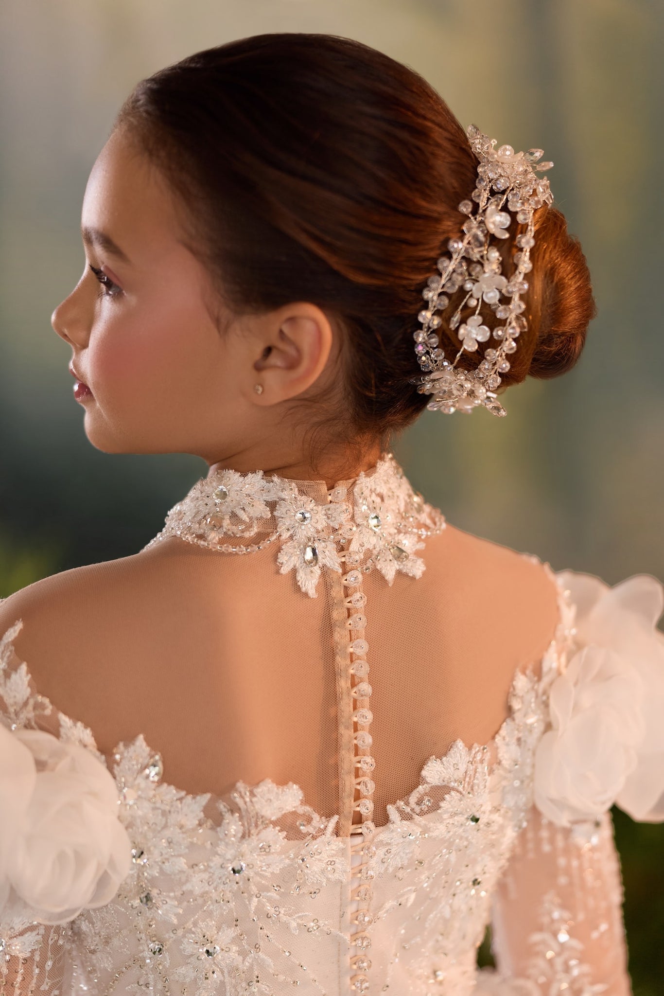 Girl wearing a white lace dress with a high neck and button back detail and a crystal hair accessory