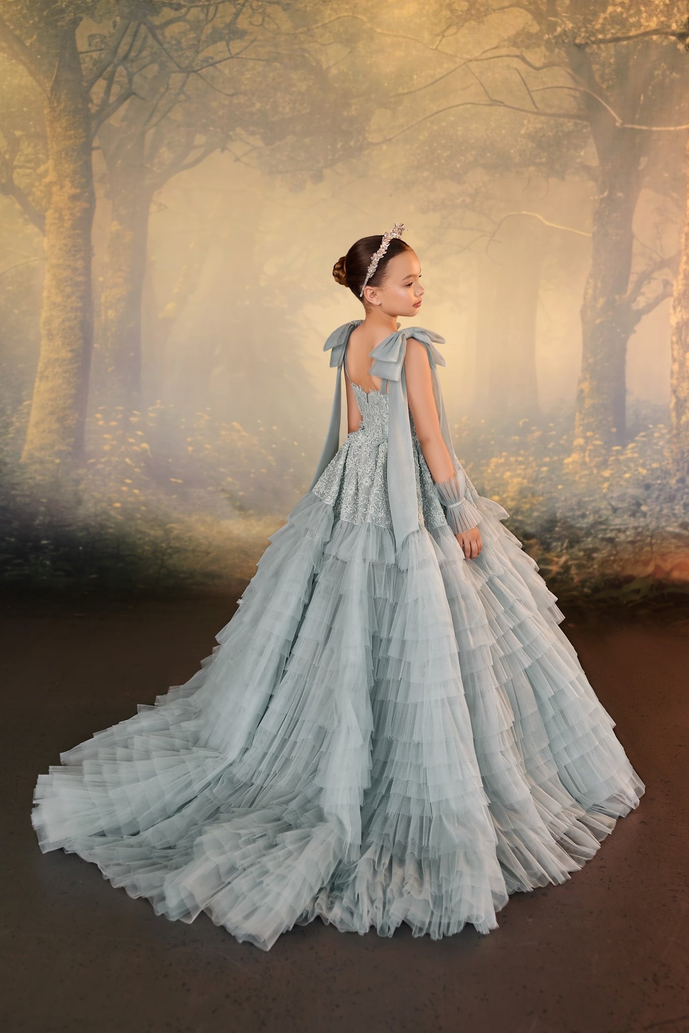 Girl wearing a dusty blue tiered tulle gown with a train and a tiara against a forest backdrop