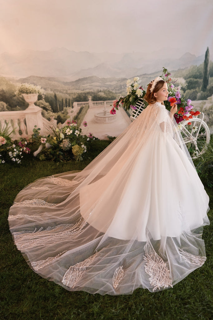 Girl wearing a white dress with a long embellished cape standing in a garden setting | Girls Formal Dress