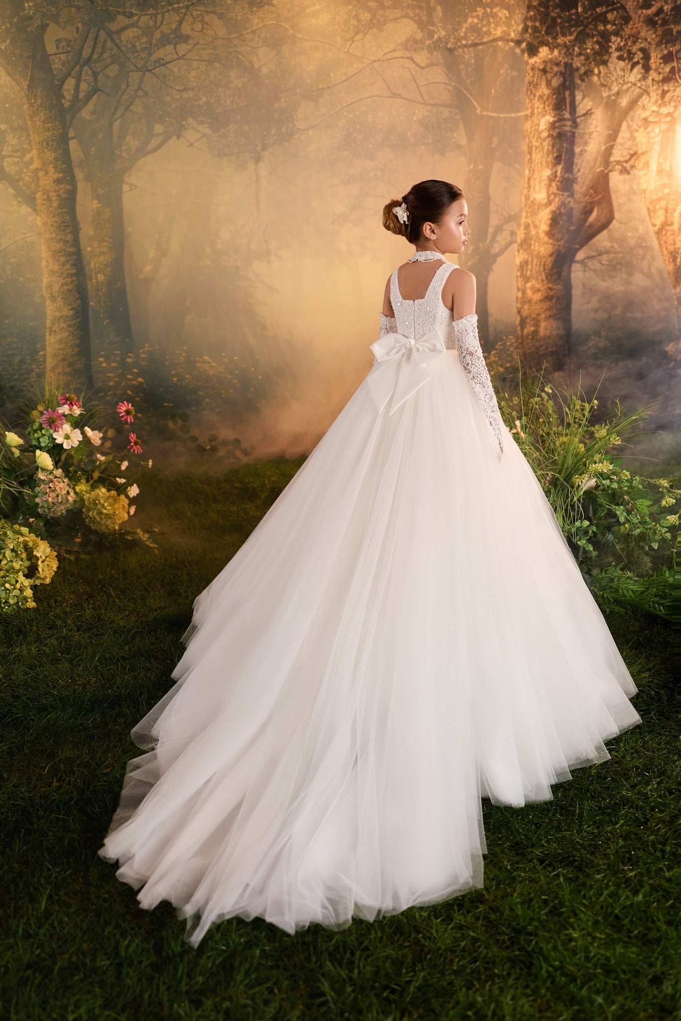 Girl wearing a white floor length tulle dress with a bow and lace gloves in a forest setting | Girls Formal Dress