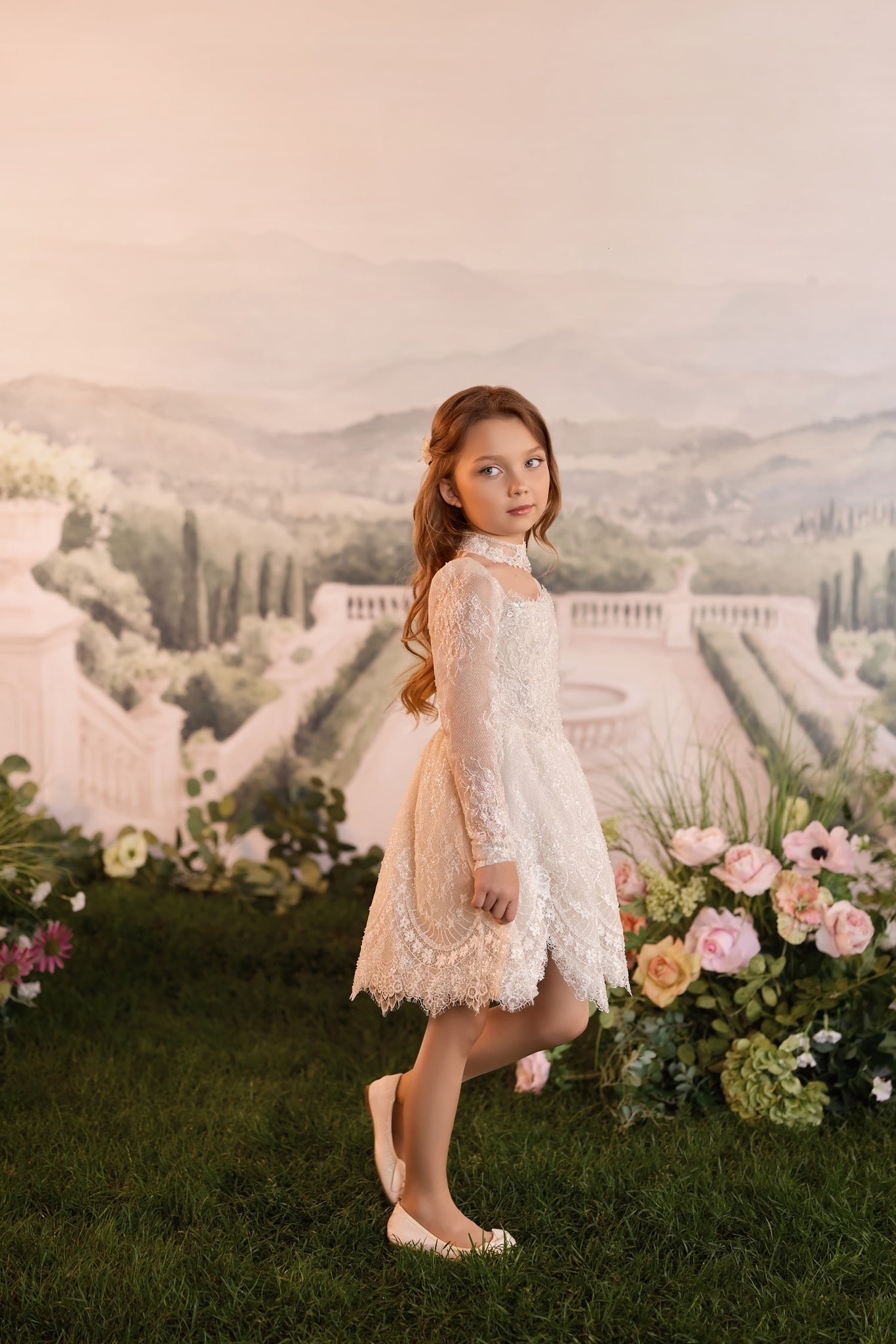A young girl wearing a light cappuccino lace dress and light cappuccino flats stands on a green lawn in front of a scenic garden backdrop with flowers and a distant landscape mural | Girls Formal Dress
