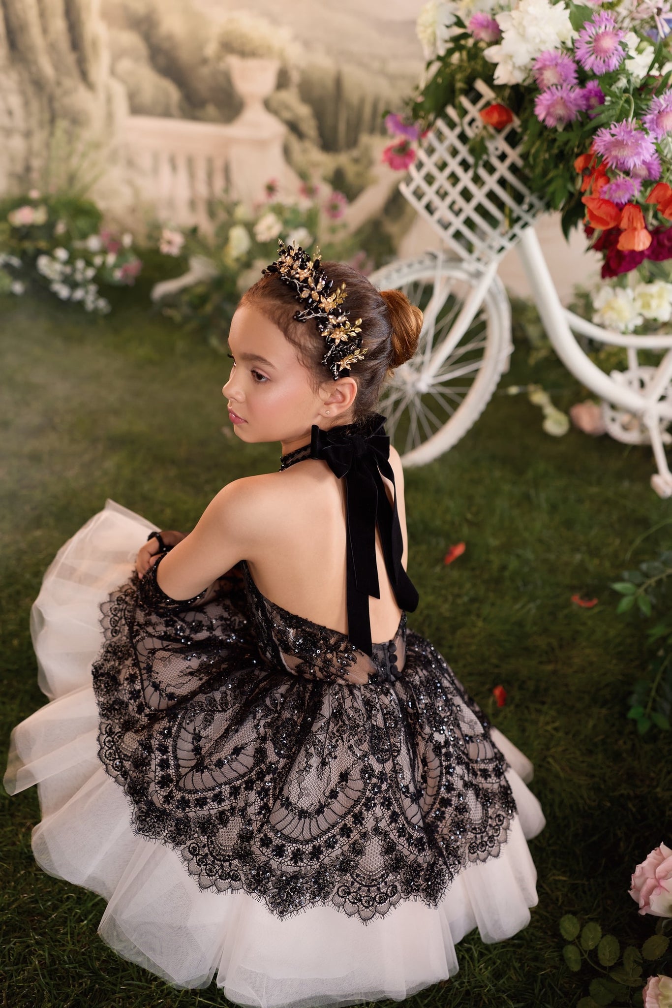 Girl wearing black lace dress and gold floral headband sitting on grass near white bicycle with flowers | Girls Formal Dress