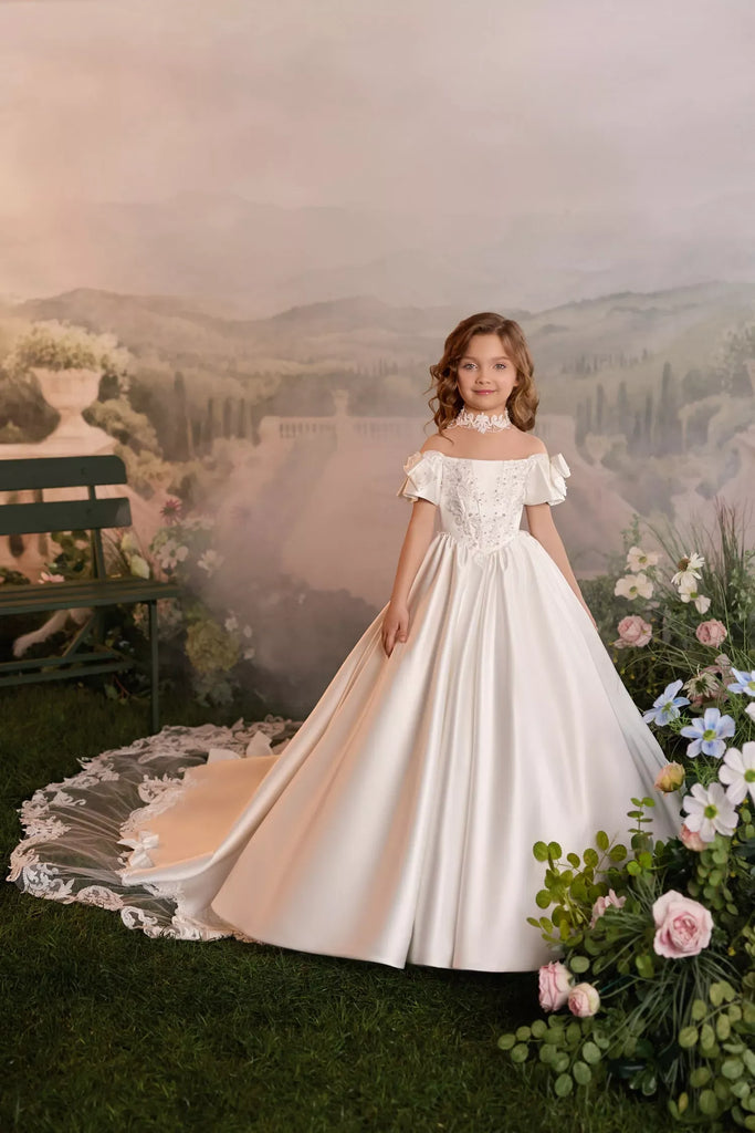 Girl wearing a white off-shoulder ball gown with lace details and a long train in a garden setting | Girls Formal Dress