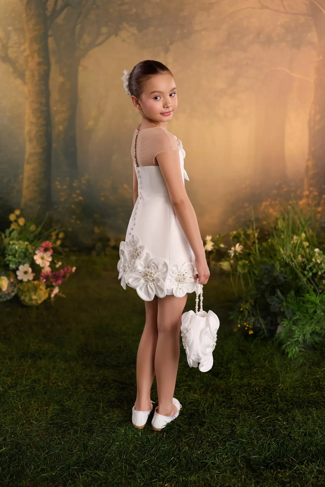 Girl wearing a white sleeveless dress with floral hem details and holding a small matching bag