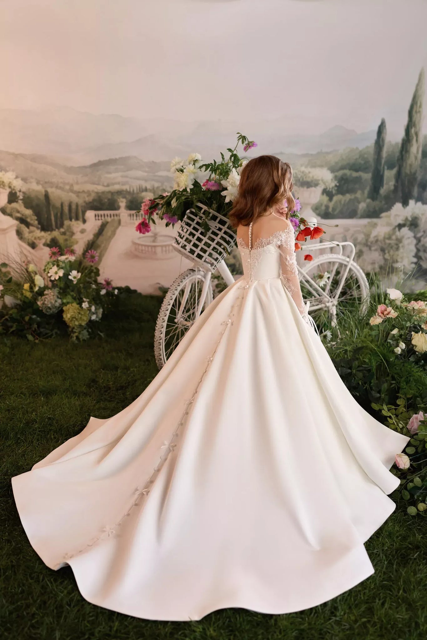 Girl wearing a white long sleeve ball gown with a train standing next to a white bicycle with a flower basket