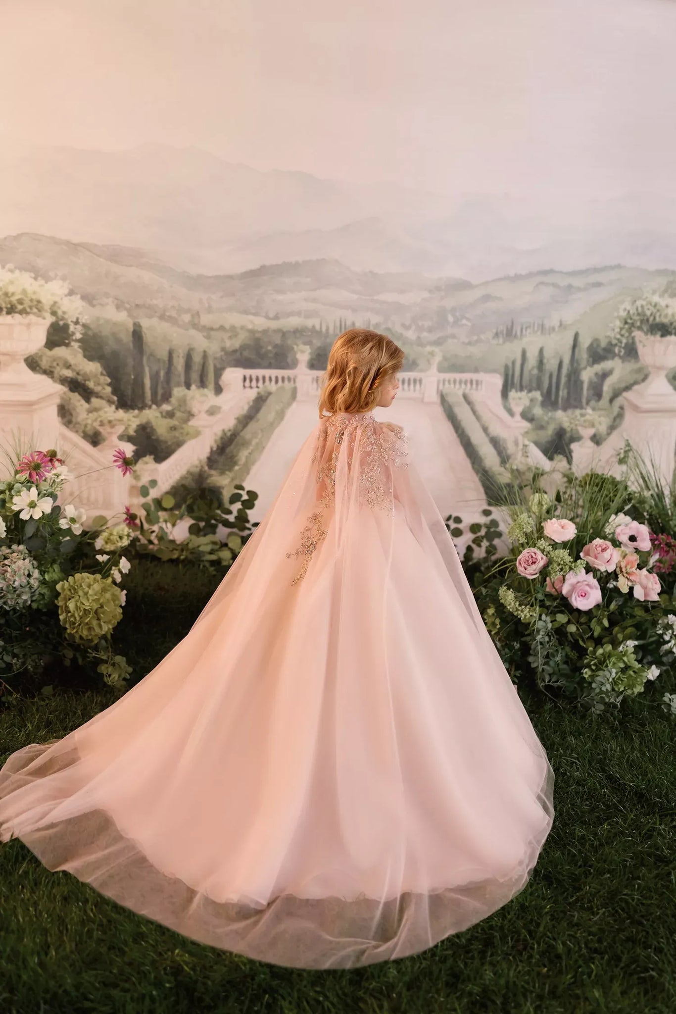 Child wearing a long pink tulle dress with lace detail standing in front of a garden backdrop