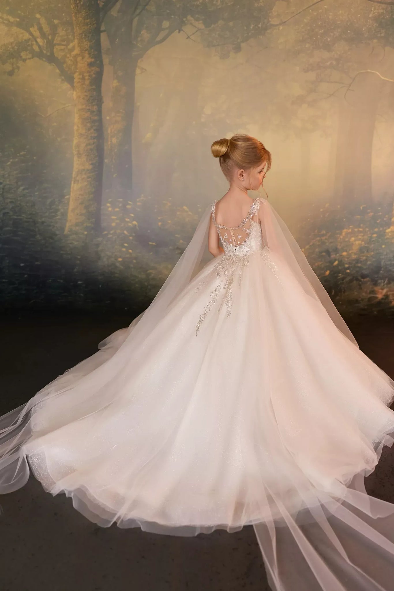 Girl wearing a white ball gown with a beaded bodice and long tulle cape against a forest backdrop