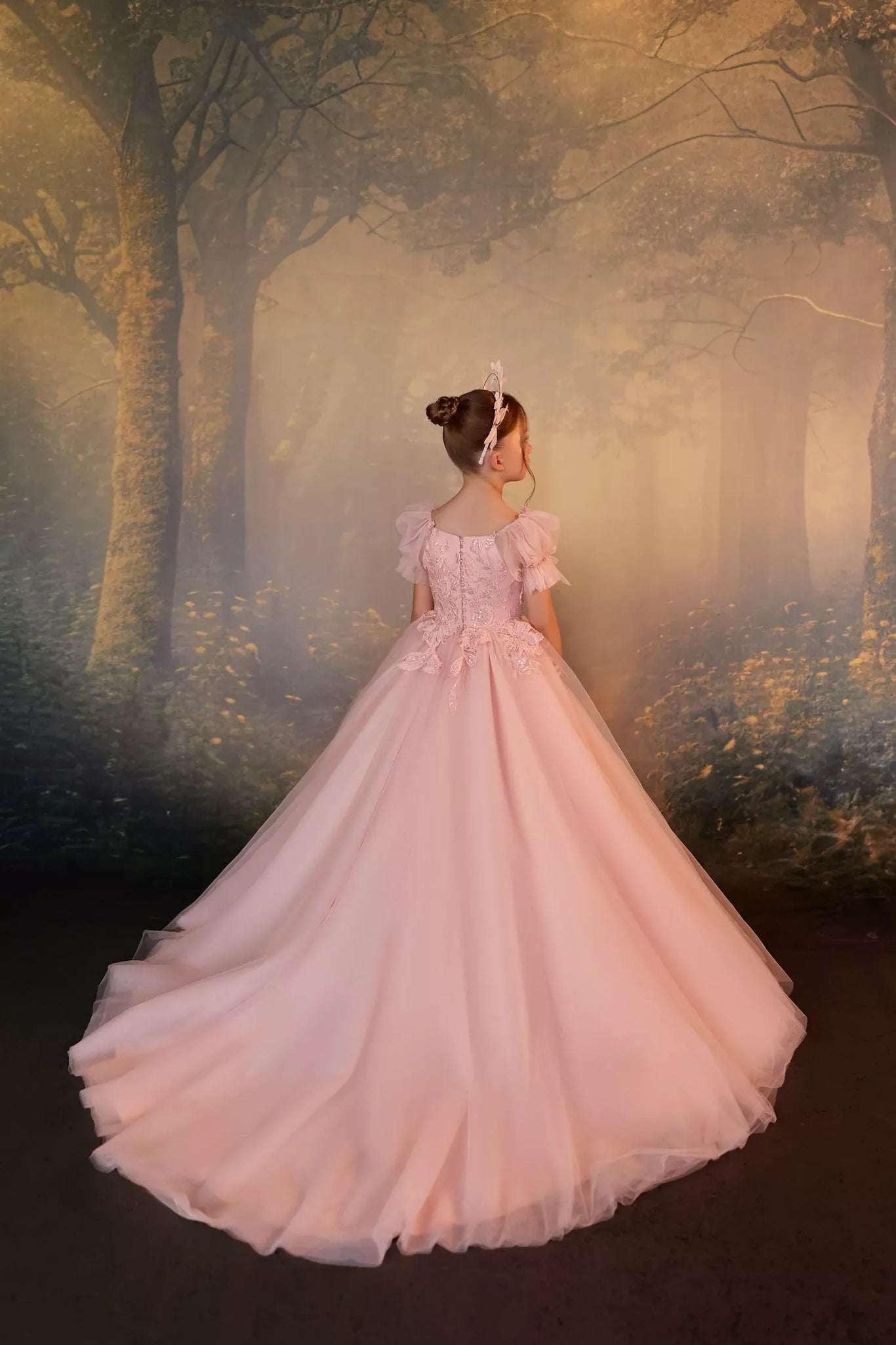 Girl wearing a pink floor length ball gown with lace bodice and puff sleeves against a forest backdrop