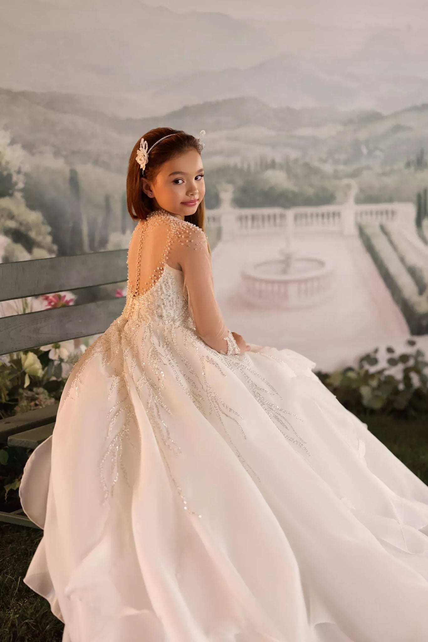 Girl wearing a white long sleeve formal dress with pearl and sequin embellishments sitting on a bench
