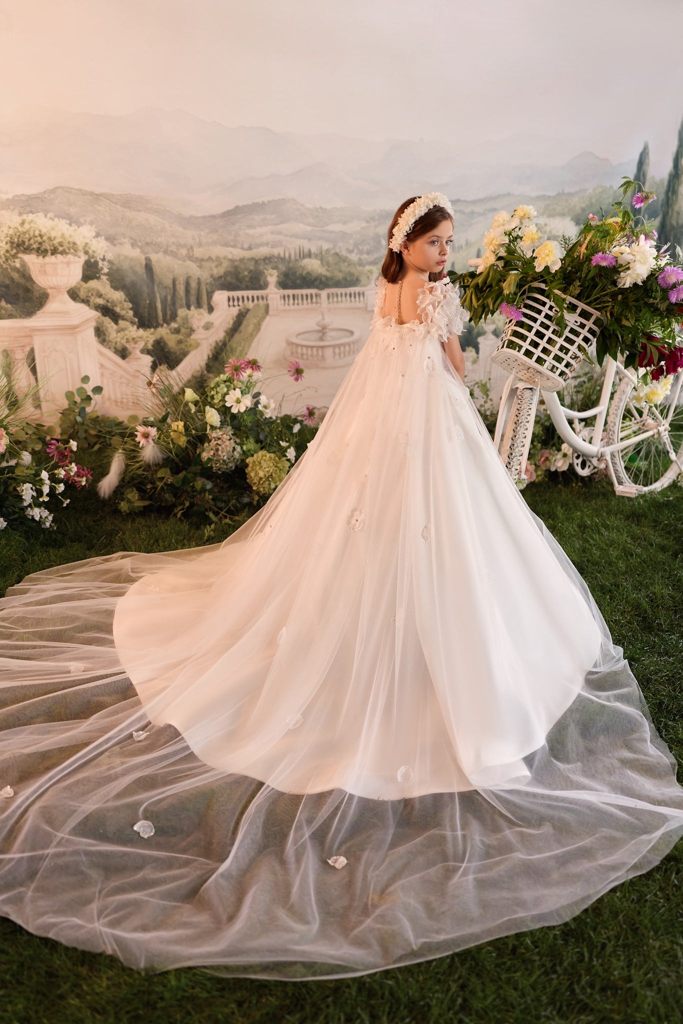 Girl in white floral dress with long tulle train standing by a white bicycle with a flower basket