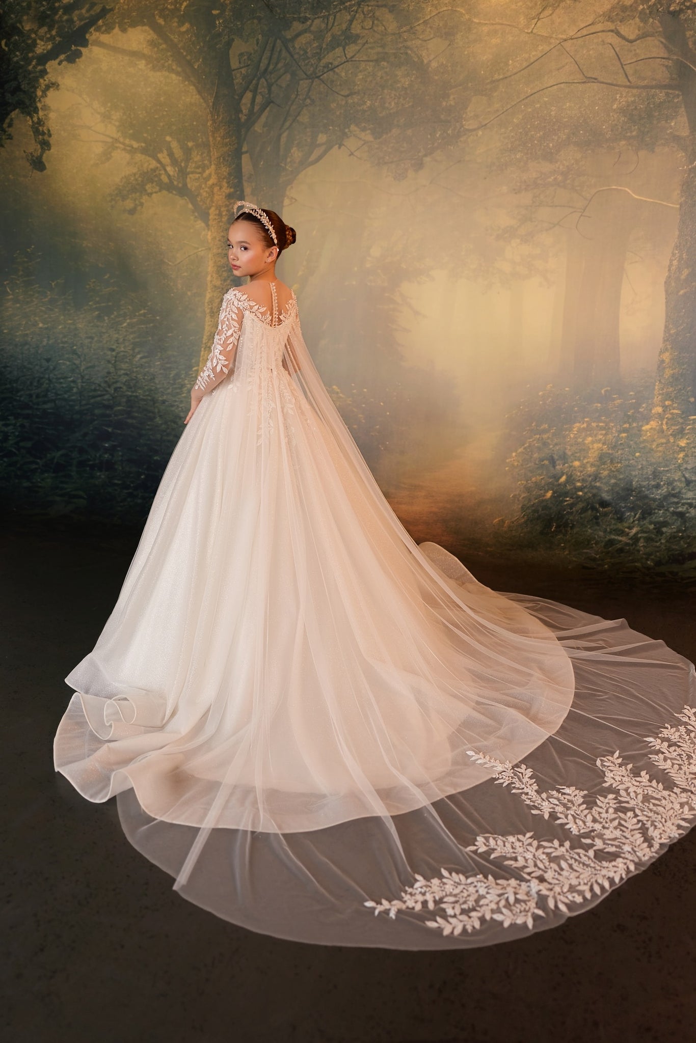 Girl wearing a long white lace gown with a train and a tiara standing in a forest setting