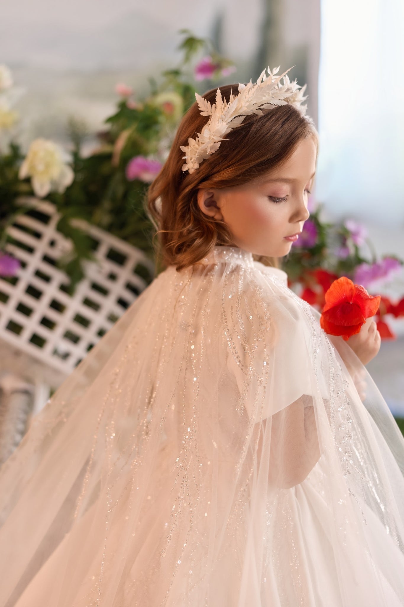 Girl wearing white sequined cape and floral headpiece holding a red flower