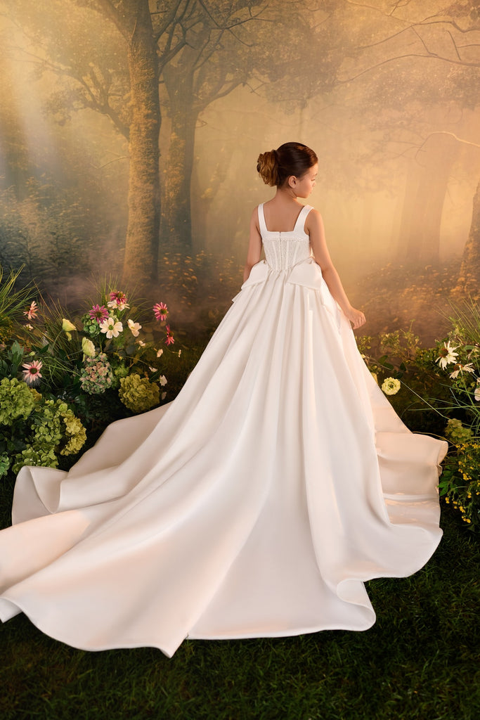 Girl wearing a white floor length dress with a train standing in a forest setting | Girls Formal Dress
