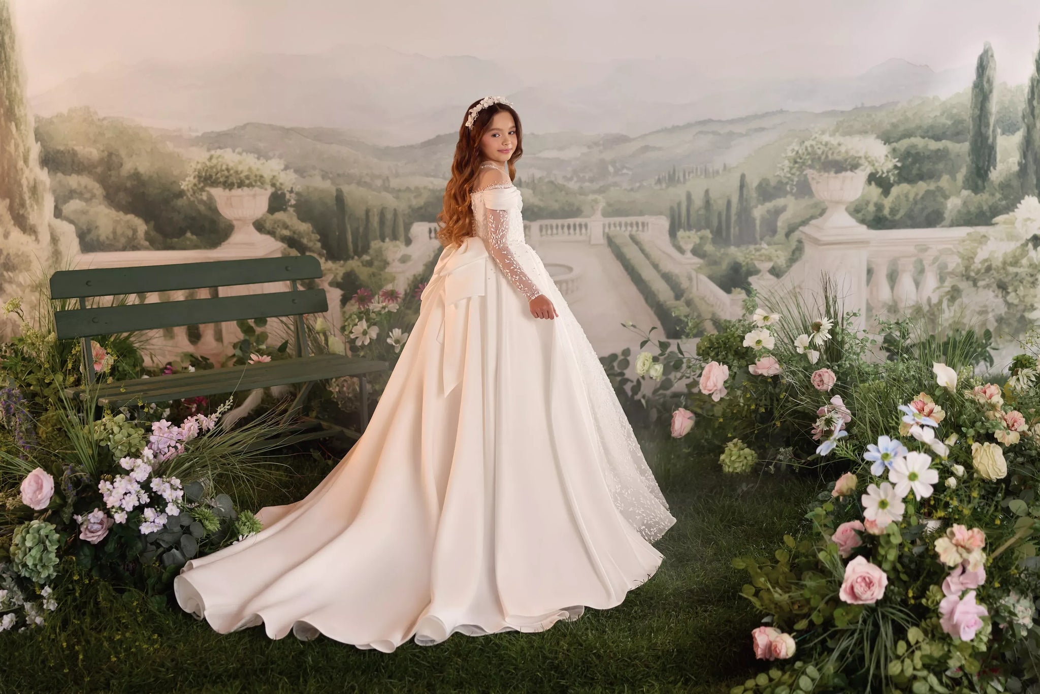Girl wearing a white off the shoulder ball gown with lace sleeves and a floral headpiece in a garden setting