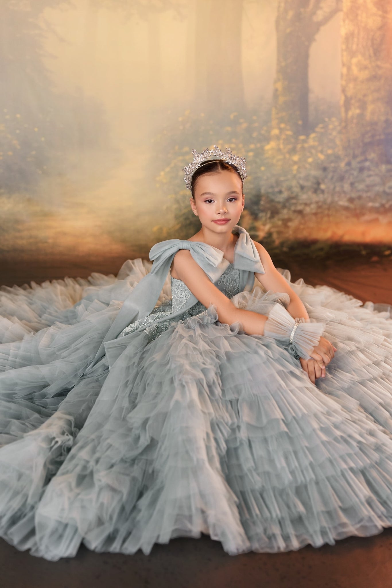 Girl wearing a grey ruffled ball gown and a silver tiara sitting against a forest backdrop