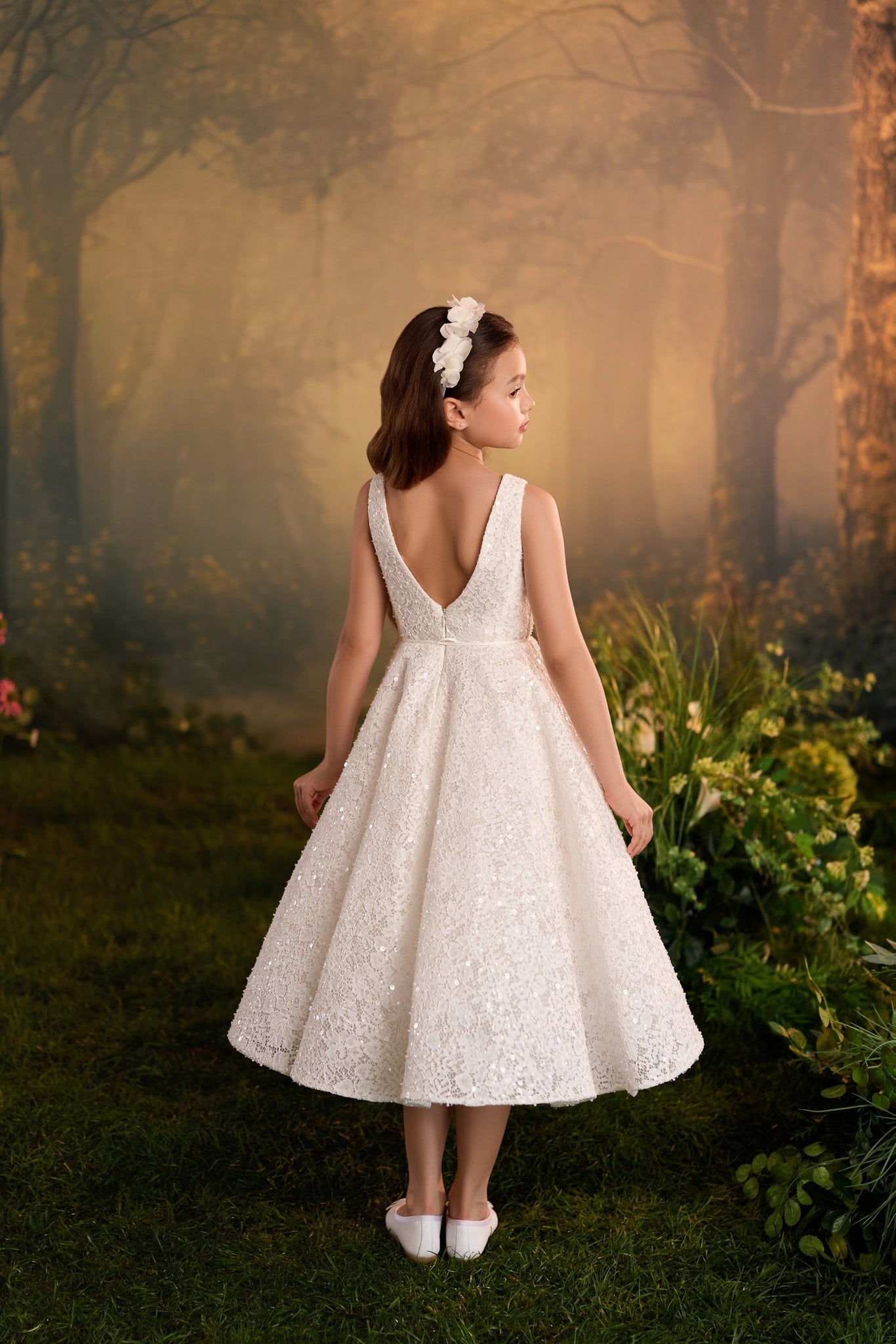 Girl wearing a white sequined A-line dress with a V-back and floral headband in a forest setting