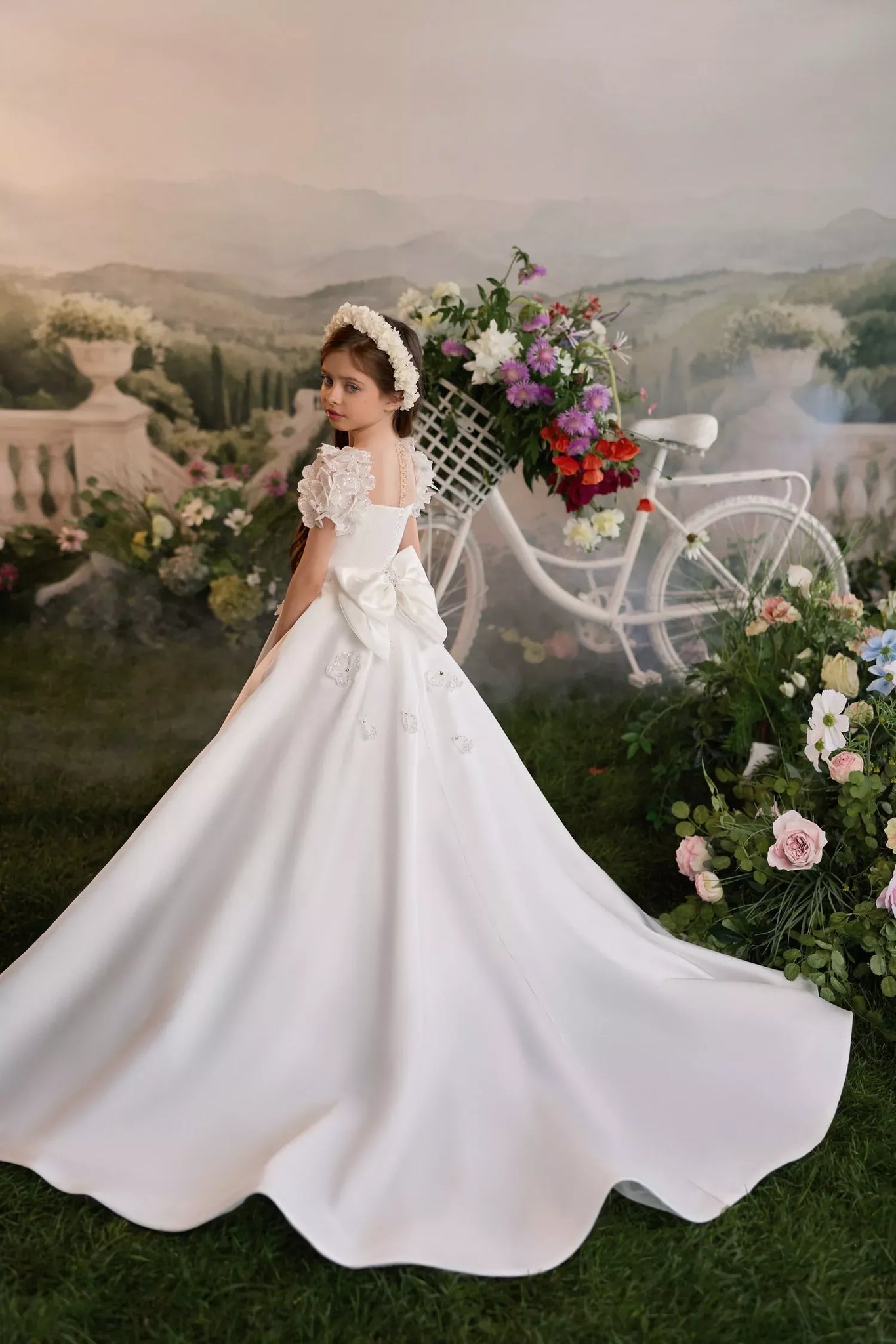 Girl wearing a white floral dress with a train and a flower crown standing by a bicycle with a basket of flowers