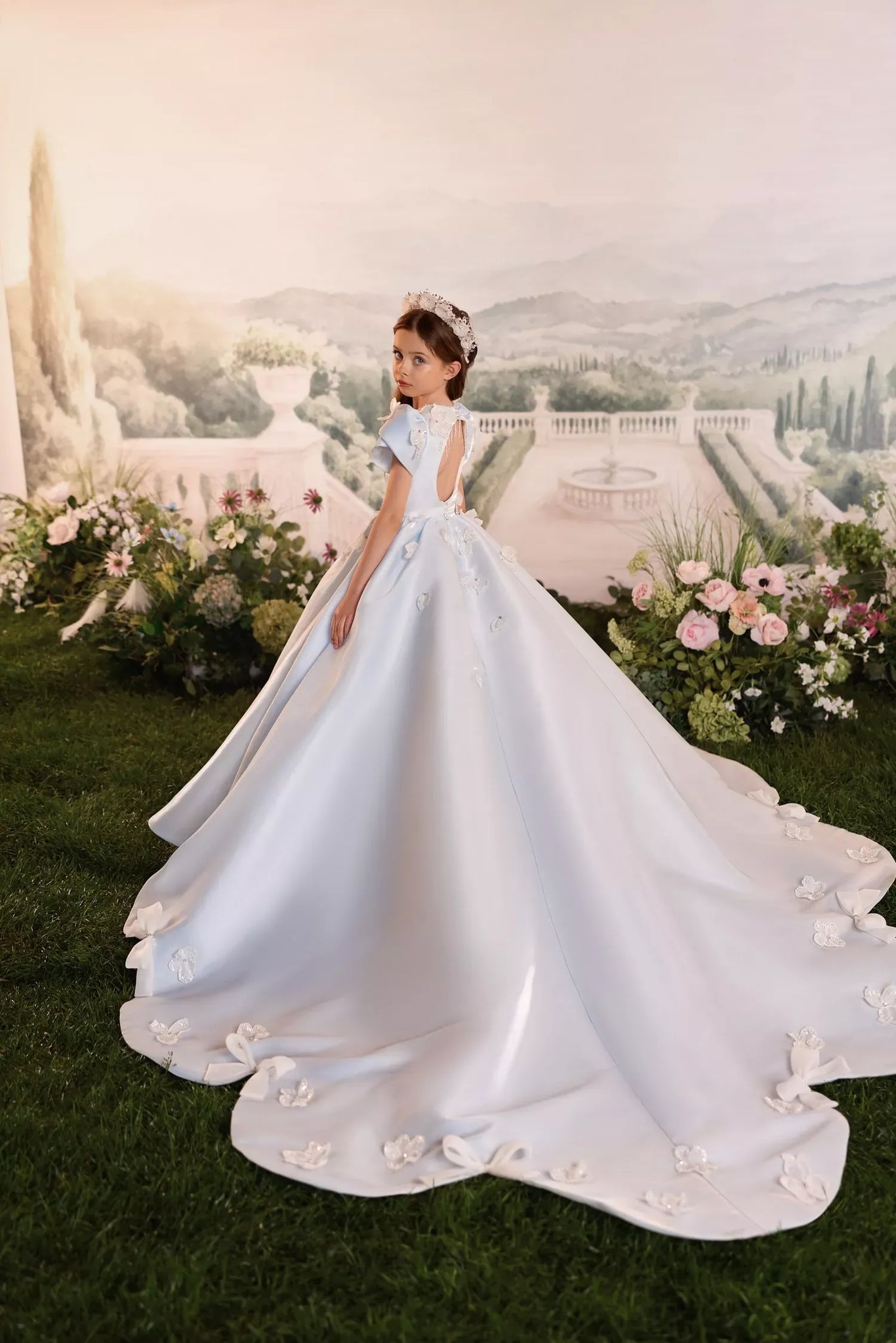 Girl wearing a light blue ball gown with a long train and floral embellishments standing in a garden setting
