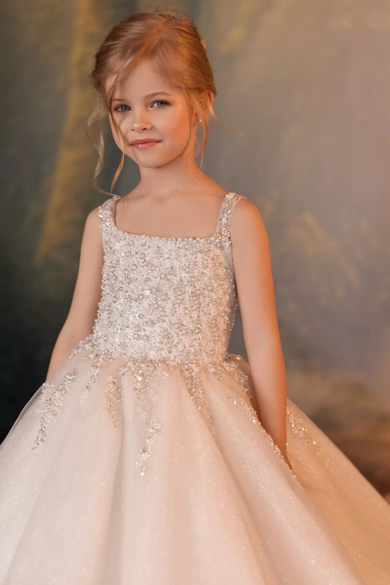 A young girl with blonde hair wearing a sparkling champagne-colored ball gown with intricate beadwork and a full skirt, posing against a soft, warm-toned studio backdrop.