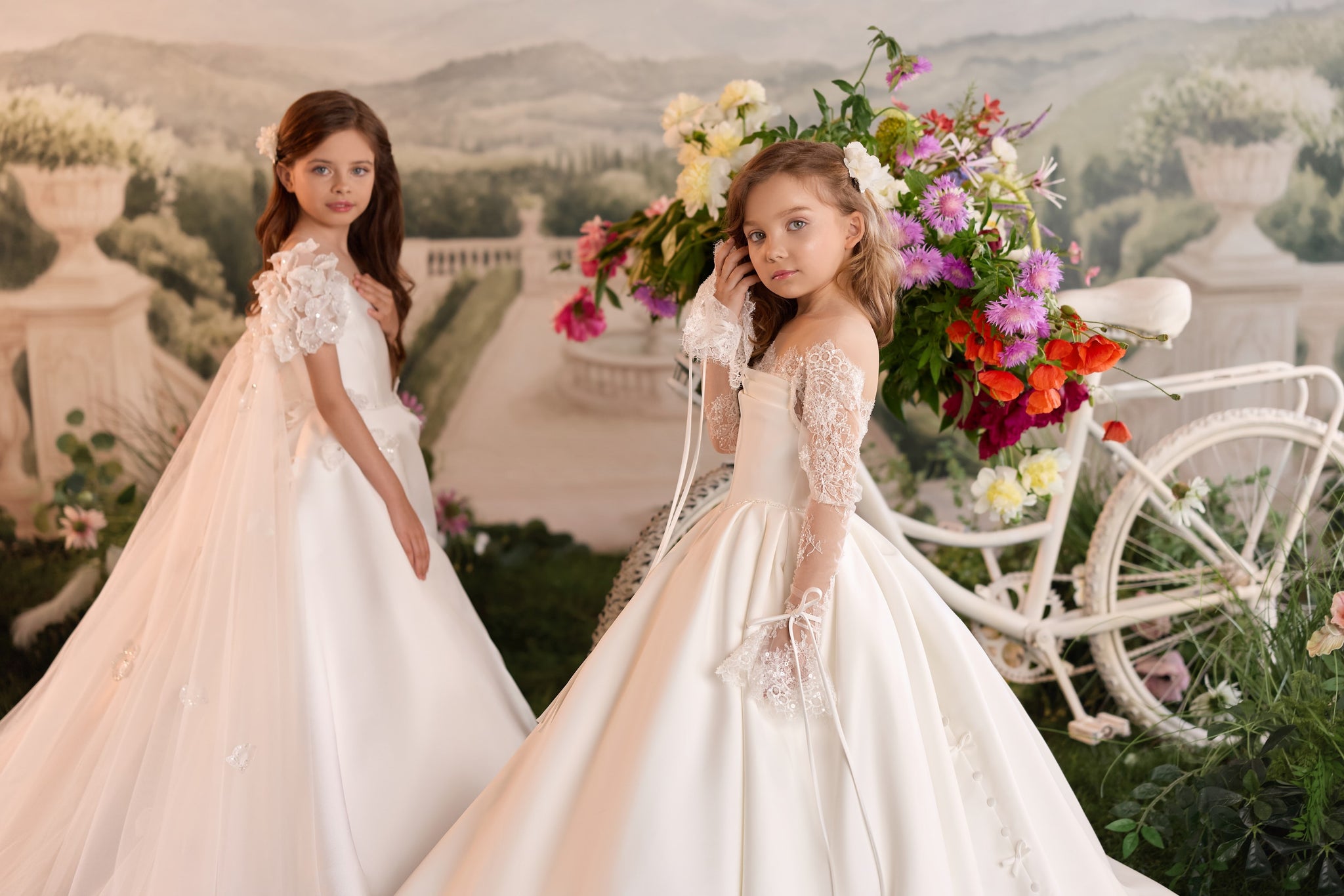 Two young girls wearing white formal dresses with lace sleeves standing near a decorative bicycle with flowers