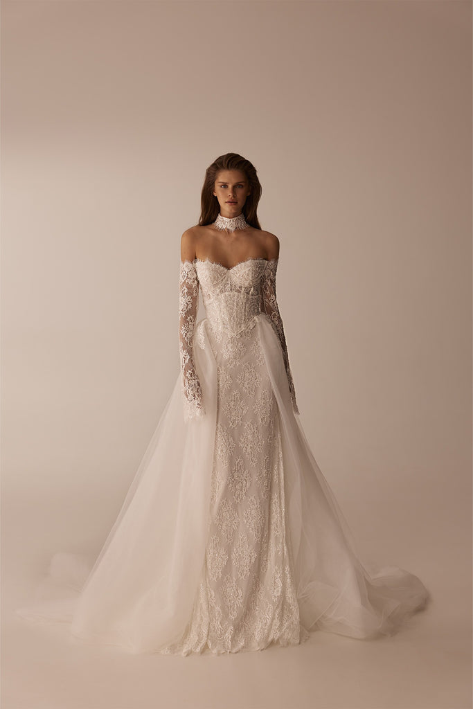 Model wearing a strapless lace wedding gown with a corset bodice, long lace sleeves, and a detachable tulle overskirt