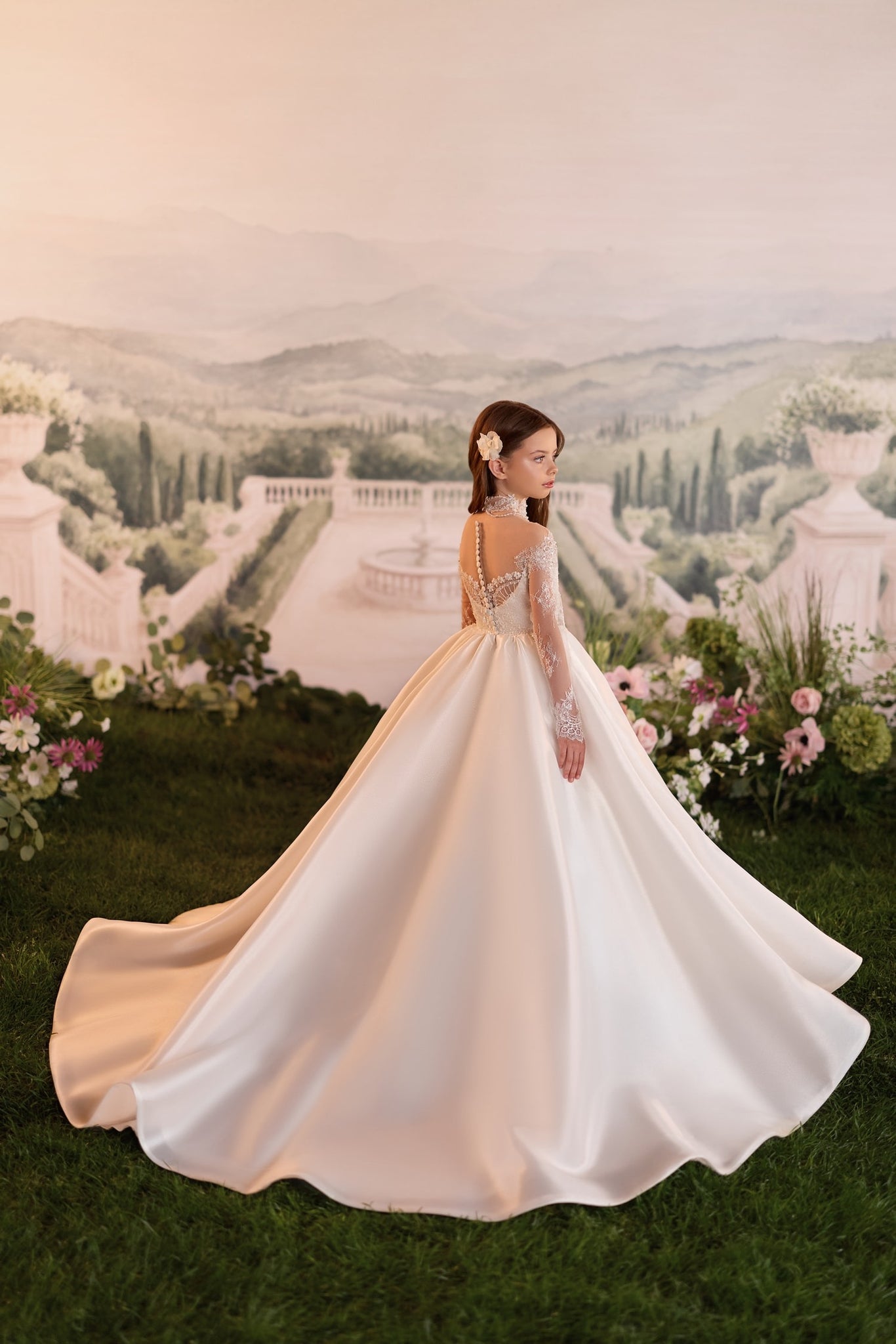 Girl wearing a long white satin dress with lace sleeves and a high neckline standing in a garden setting