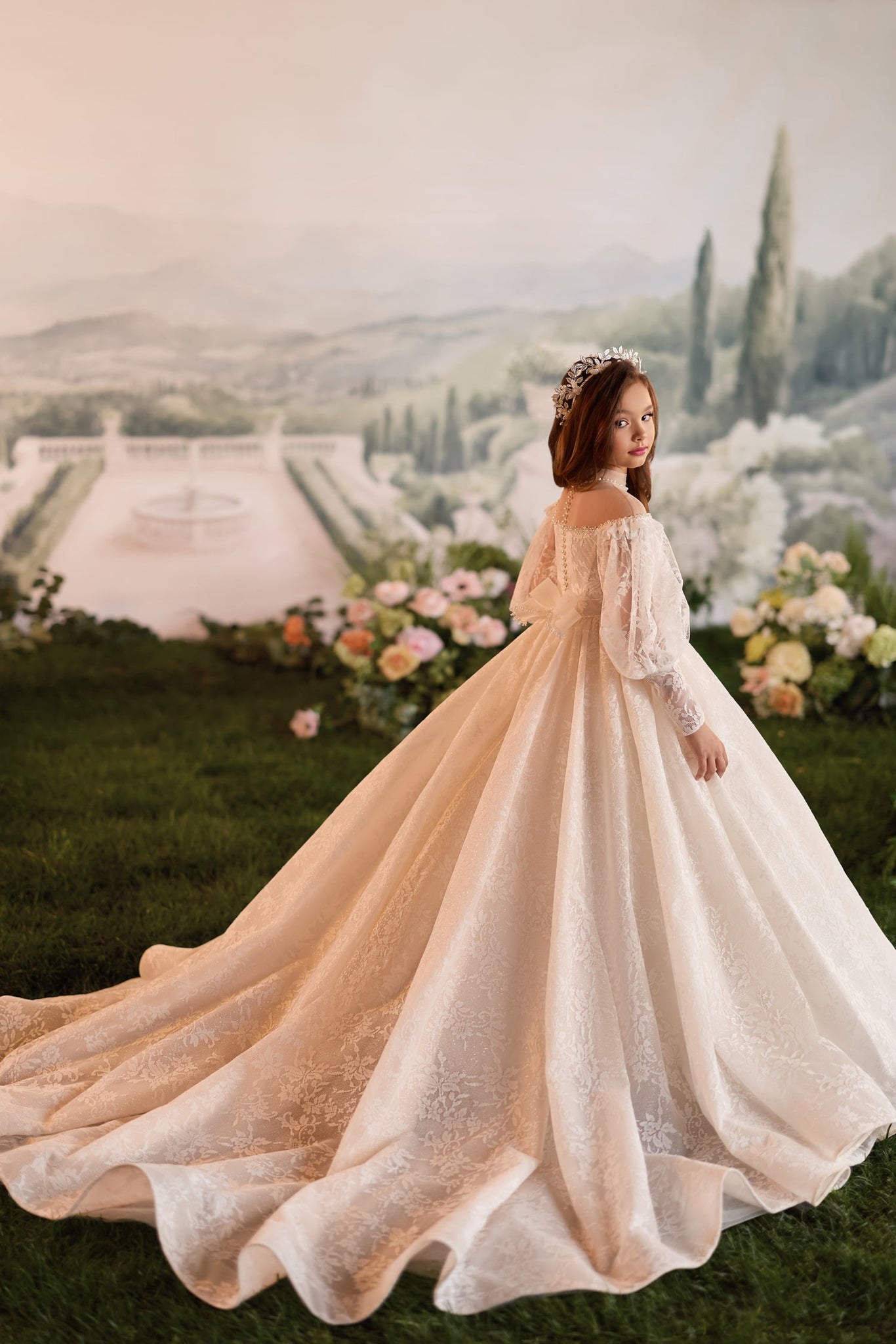 Girl wearing a white lace ball gown with long sleeves and a tiara standing in a garden setting