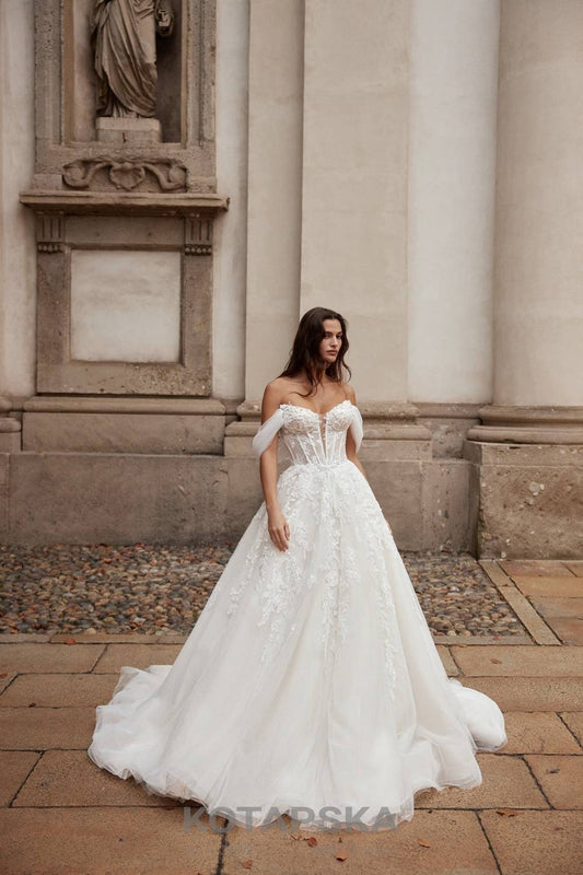 Model wearing a lace ball gown wedding dress with corset bodice, off-shoulder tulle sleeves, and full skirt in an elegant courtyard