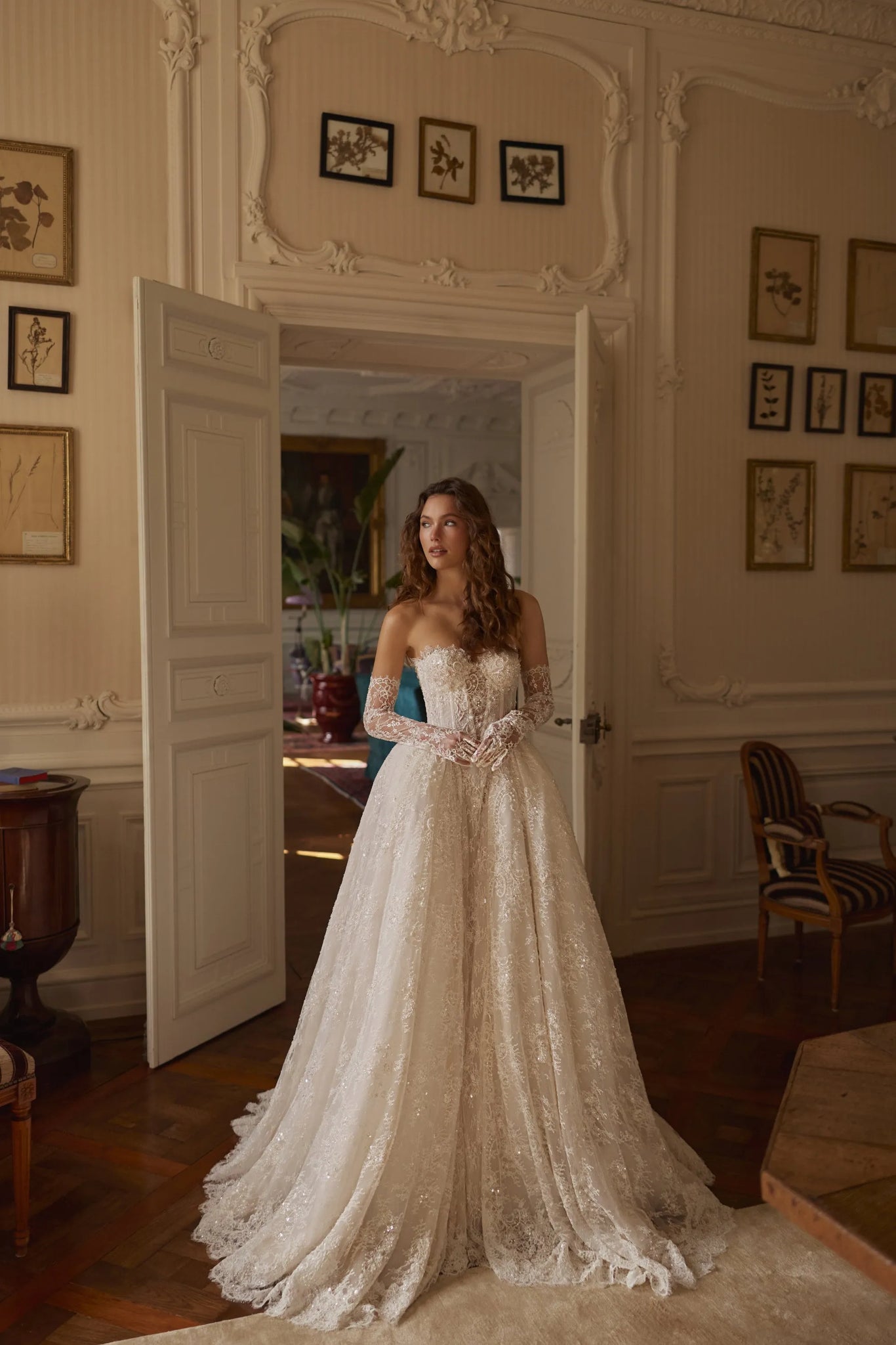 A woman wearing a voluminous ivory lace wedding gown with long sheer sleeves stands in an ornate room with cream walls, framed botanical prints, and a large open doorway behind her.