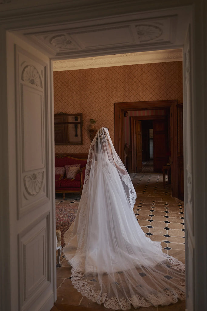 Bride in a long white lace wedding dress and veil standing in a room viewed through a doorway