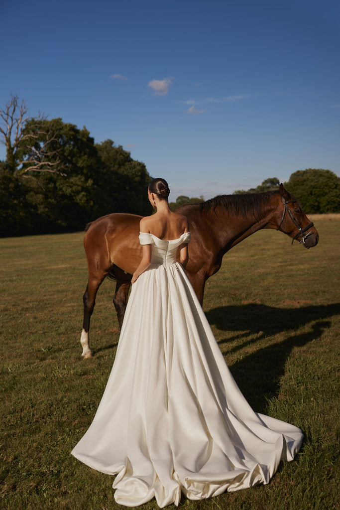 Woman in white off-shoulder wedding dress standing in a field next to a brown horse
